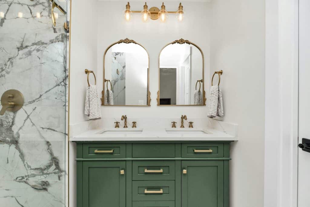 Bathroom remodel with a green vanity, brass fixtures, arched mirrors and a marble-tiled shower